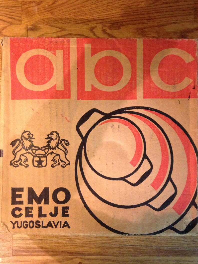 Puede incluir: Una caja de cart&oacute;n vintage con las letras "ABC" en un rect&aacute;ngulo rojo en la parte superior. Debajo, la caja presenta un gr&aacute;fico de una olla con detalles en rojo y el texto "EMO CELJE YUGOSLAVIA". La caja tiene un escudo de le&oacute;n.