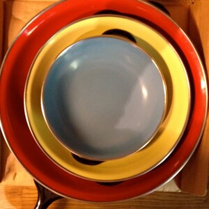 Puede incluir: Un conjunto anidado de utensilios de cocina coloridos. Una sart&eacute;n roja forma la base, seguida de un plato amarillo y rematada con un plato azul claro. Los utensilios de cocina tienen bordes plateados y un asa negra.