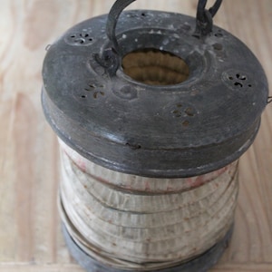 Puede incluir: Linterna de metal antiguo con tapa y base circulares, y una pantalla cilíndrica de papel. La parte superior tiene recortes decorativos y un asa para colgar. La linterna tiene un aspecto envejecido, lo que sugiere que es antigua o una réplica.