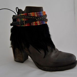 May include: Brown leather ankle boot with a block heel, adorned with black feathers, a colorful woven band, and studded black leather straps. The boot has a buckle closure and a rounded toe.