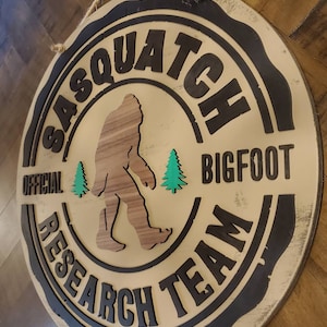 May include: A round wooden sign with a black border and the text "Sasquatch Bigfoot Research Team" in black lettering. The sign features a brown silhouette of a Sasquatch walking with two green pine trees.