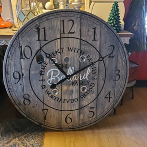 May include: A wooden clock with a distressed gray finish. The clock face features black Roman numerals and a black hand. The clock is decorated with the text "Time spent with family is worth every second" and "The Ballard Family".