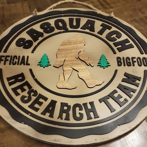 May include: A round wooden sign with a black border and the text "Official Sasquatch Bigfoot Research Team" in white lettering. A silhouette of Bigfoot is in the center of the sign, with two green trees on either side.