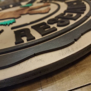 May include: A wooden sign with a black border and the words "Trespassers Will Be Resented" in black lettering. The sign features a cartoon image of a person with a green bush on their head and legs.