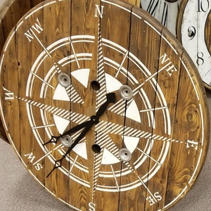 May include: A wooden clock with a compass design. The clock face is painted white with black markings and a black clock hand. The clock is mounted on a wooden spool.
