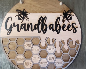May include: A wooden sign with the text "Grandbabees" in black lettering. The sign is decorated with a honeycomb pattern and has the names of grandchildren written in the honeycomb cells. Two black silhouette bees are on either side of the text.