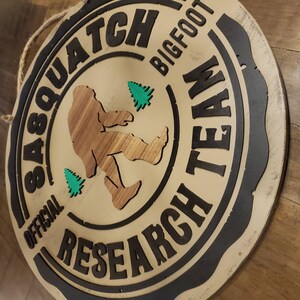 May include: A round wooden sign with a black border and the text "Official Sasquatch Research Team Bigfoot" in white lettering. A brown silhouette of a sasquatch is walking with two green trees in the background.