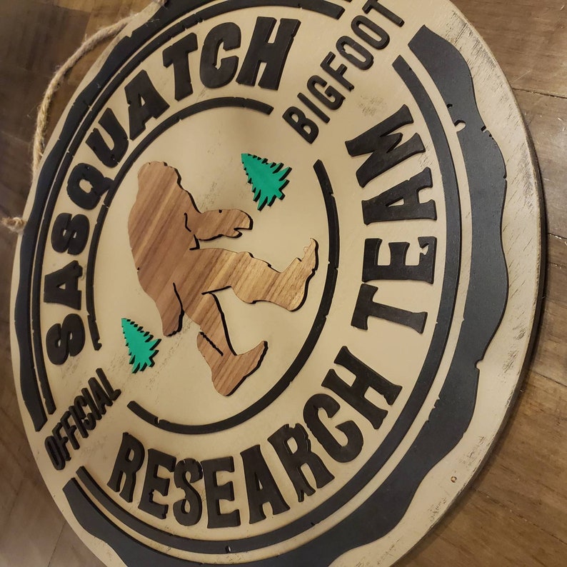May include: A round wooden sign with a black border and the text "Official Sasquatch Research Team Bigfoot" in white lettering. A brown silhouette of a sasquatch is walking with two green trees in the background.