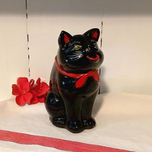 Könnte beinhalten: Glänzende schwarze Katzenfigur mit rotem Band um den Hals und roten Akzenten an den Ohren. Die Katze hat grüne Augen und einen lächelnden Ausdruck. Rote Blumen liegen neben der Katze.