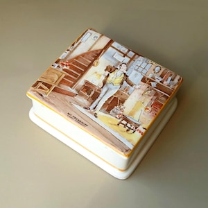May include: A white ceramic box with a yellow trim and a colorful illustration of a man in a room with a fireplace and a staircase. The box has the text "Mr. Pecksniff (London, 1837)" on the bottom.