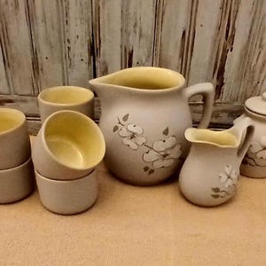 May include: A set of vintage ceramic kitchenware with a floral design. The set includes a pitcher, a creamer, a sugar bowl, and four cups. The pieces are all in shades of beige and brown with a yellow glaze on the inside.