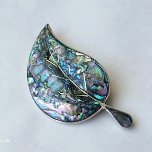 May include: A leaf-shaped brooch with iridescent abalone shell inlay. The brooch has a silver-tone metal frame and features a detailed veining pattern. The abalone shell displays a range of colors, including blue, green, and purple.