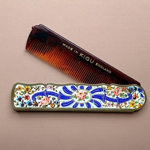 May include: A brown tortoiseshell-style plastic comb with the text MADE IN KIGU ENGLAND printed in gold. The comb is partially inserted into a rectangular metal case featuring a decorative enamel pattern of colorful flowers and blue ribbons.