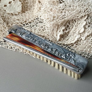 May include: A rectangular grooming tool featuring a silver-toned metal handle with an embossed landscape scene of trees and buildings. The tool includes a row of off-white bristles and a brown, translucent, fine-toothed comb integrated into the side.