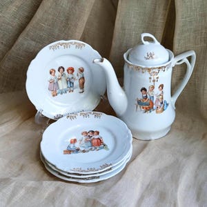 May include: A white porcelain tea set with a teapot and several small plates. Each piece features a decorative gold trim and illustrations of children in colorful clothing. The teapot has a curved spout and a handle.