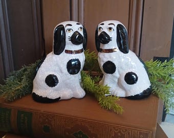 May include: Two ceramic dog figurines. The dogs are white with black spots, ears, and noses. They have brown collars and are sitting on a vintage book. The background is a dark wooden surface.