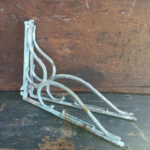 May include: Two antique, weathered, cast iron shelf brackets with a distressed, light blue-green finish. The brackets have an ornate, curved design and are suitable for supporting shelves. The brackets are displayed on a dark, textured surface.