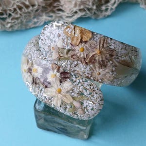 May include: White resin bangle bracelet with embedded dried flowers and white glitter. The flowers are white, brown, and yellow.