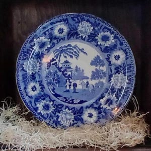 May include: A blue and white decorative plate with a floral border and a central landscape scene. The plate is displayed on a bed of white decorative filler, showcasing a classic design.
