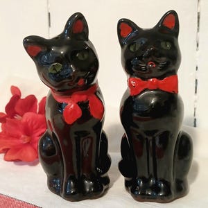 Könnte beinhalten: Zwei schwarze Katzenfiguren mit roten Akzenten. Eine Katze hat eine rote Fliege, die andere einen roten Schal. Beide haben grüne Augen und sitzen aufrecht. Im Hintergrund eine rote Blume.