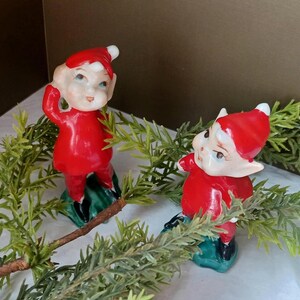 May include: Two vintage-style elf figurines wearing bright red outfits with matching pointed hats and black shoes. Each elf has a pale complexion, pointed ears, and is positioned standing on a small green base, surrounded by green pine branches.