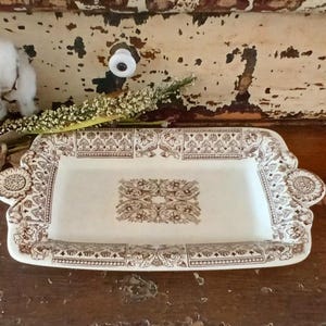 May include: A rectangular ceramic tray featuring a cream-colored base with intricate brown floral and geometric patterns. The tray has a decorative border and two circular handles on the sides, each adorned with a detailed brown circular motif.