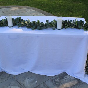 Puede incluir: Un mantel blanco con una guirnalda de eucalipto y dos velas blancas en portavelas de vidrio. La mesa está preparada al aire libre en un patio.