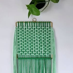 Może przedstawiać: Wisząca ozdoba ścienna z makramu w kolorze zielonym i jasnozielonym z tkanym wzorem i frędzlami. Ozdoba jest oprawiona w złotą metalową ramę.