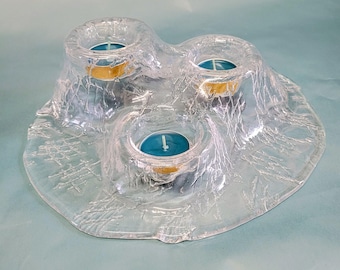 May include: A clear glass candle holder with three tealight candle holders. The holder has a textured, frosted appearance and a unique, organic shape. The tealight candles have gold-colored rims.