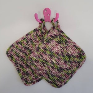 Könnte beinhalten: Zwei gehäkelte Topflappen in Grün-, Rosa- und Weißtönen, die an einem rosa Haken hängen.