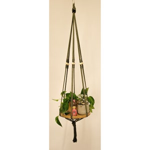 May include: A green macrame plant hanger with wooden beads and a wooden shelf. The hanger is holding two small potted plants.