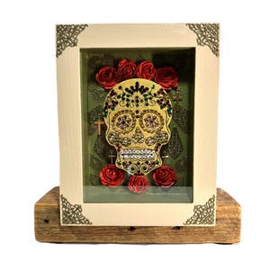 Könnte beinhalten: Ein Schattenbox-Rahmen mit cremefarbenem Rand und verzierten Ecken enthält einen goldfarbenen Totenkopf, der mit bunten Strasssteinen besetzt ist. Der Totenkopf ist von roten Papierrosen und kleinen goldenen Kreuzen auf grünem Grund umgeben.