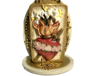 May include: A decorative plaque featuring a central relief of a red heart adorned with pink roses, pierced by a dagger, and topped with flames and white lilies. The rectangular piece is framed by gold beads and small crosses on an oval base.