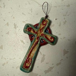 May include: A ceramic cross ornament featuring a glossy finish with a teal border, a cream-colored center, and raised red swirl patterns. The cross has a circular element behind the intersection and includes a thin dark wire loop for hanging.