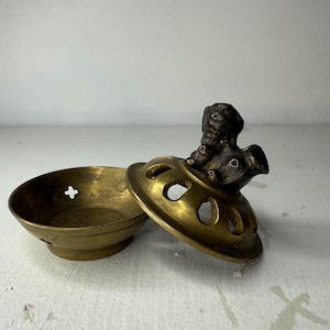 May include: A vintage brass incense burner with a detailed lid featuring a small, ornate figure. The burner has a bowl-like base and a perforated lid with a decorative design. The brass has a warm, aged patina.