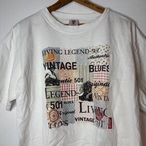 May include: A white short-sleeve t-shirt featuring a central graphic print composed of various patchwork-style squares with patterns and text including "LIVING LEGEND," "VINTAGE," "BLUES," "501," "Authentic," and "Original" in black and muted colors.