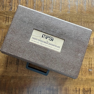 May include: A rectangular brown textured plastic carrying case with a black handle. A central white label features the text MPSI, MICRO PROCESSOR SYSTEMS, INC., 7641 19 MILE ROAD, STERLING HEIGHTS, MI 48078, and the phone number (313) 731-6410.