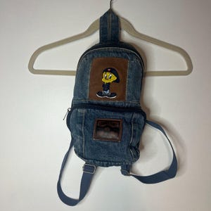 May include: A blue denim backpack with a brown leather patch featuring a cartoon Tweety Bird wearing a red cap. The backpack has a zipper closure and adjustable straps.
