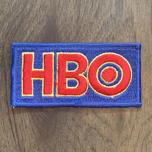 May include: A rectangular embroidered patch featuring the letters HBO in bold red thread with a thin yellow outline. The background of the patch is a solid blue color, and the entire piece is finished with a matching blue stitched border.