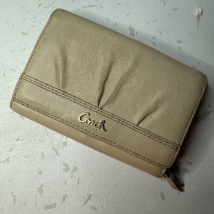 May include: A beige leather wallet featuring a textured surface with subtle pleating details across the top section. The front displays the gold-toned Coach logo in a script font, positioned above a horizontal stitched band with a zipper pull tab.