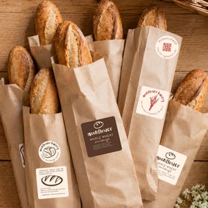 May include: A selection of sourdough loaves in brown paper bags. Each bag features a sticker with the bakery's logo and text. The bread has a golden-brown crust and is partially visible. The bags are arranged on a wooden surface.