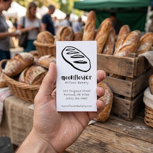 May include: A business card for Moonflower Artisan Bakery is held in front of a display of fresh bread. The card features a bread loaf illustration and the bakery's name, address, and phone number. Baskets and wooden crates hold various loaves.