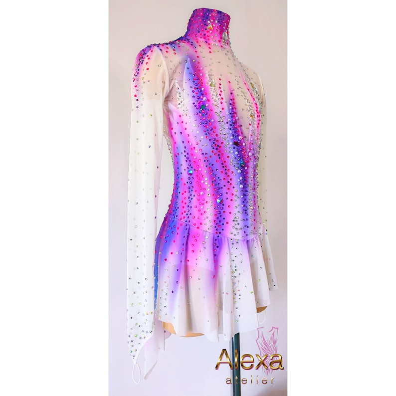 May include: A white figure skating dress with long sleeves and a high neck. The dress has a purple and pink ombre design with rhinestones throughout. The dress has a short skirt with a ruffled hem.