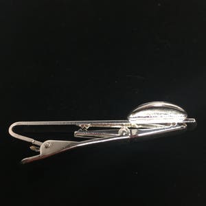 May include: Silver tie clip with a rounded, textured top. The clip is designed to hold a tie in place.