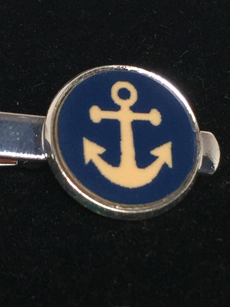 May include: A silver tie clip with a round blue enamel charm featuring a yellow anchor.