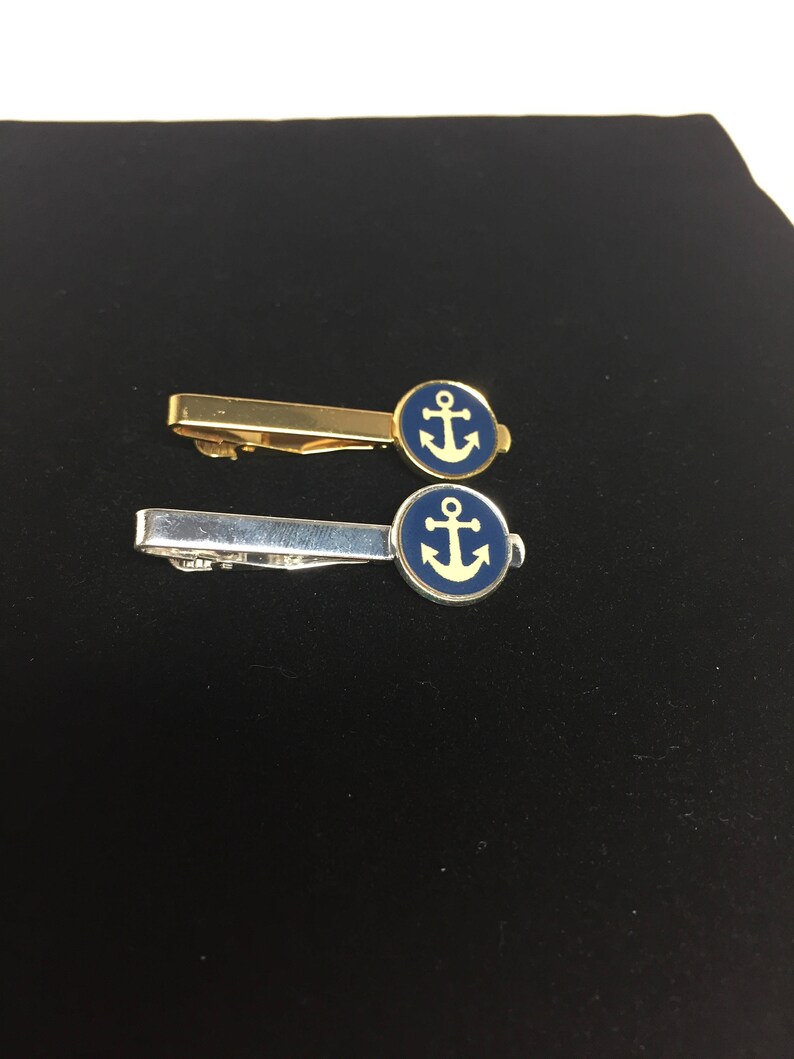 May include: Two tie clips with an anchor design. The top clip is gold with a blue enamel anchor. The bottom clip is silver with a blue enamel anchor.