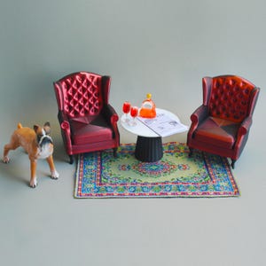 May include: Miniature living room scene with two red armchairs, a small round table, and a patterned rug. A toy dog stands near the chairs. The table holds two glasses of red liquid and a bottle.