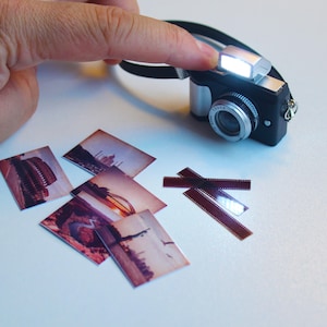 Könnte beinhalten: Eine kleine schwarze und silberne Kamera mit hellem Blitz wird von einer Hand gehalten. Mehrere kleine Fotos und Filmstreifen sind auf einer weißen Oberfläche verstreut, was auf ein Fotografie-Thema hindeutet.