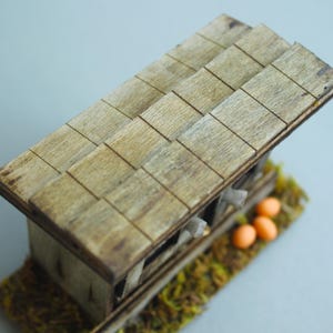 K&ouml;nnte beinhalten: Ein Miniatur-H&uuml;hnerstall aus Holz mit einem Schindeldach. Der Stall besteht aus hellbraunem Holz und steht auf einem Bett aus gr&uuml;nem und gelbem Moos. Drei orangefarbene Eier sind in der N&auml;he des Stalls zu sehen.