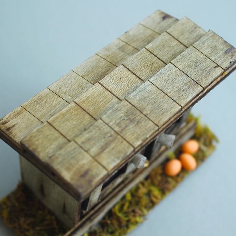 K&ouml;nnte beinhalten: Ein Miniatur-H&uuml;hnerstall aus Holz mit einem Schindeldach. Der Stall besteht aus hellbraunem Holz und steht auf einem Bett aus gr&uuml;nem und gelbem Moos. Drei orangefarbene Eier sind in der N&auml;he des Stalls zu sehen.